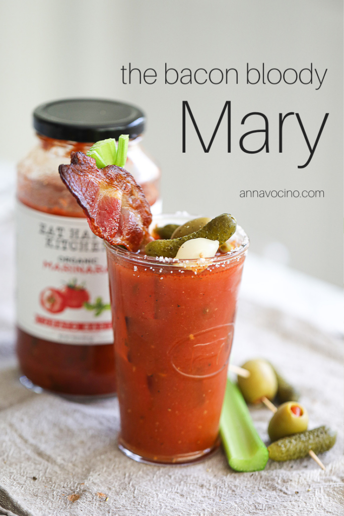 this is a PIN for Pinterest of the the bacon Bloody Mary with celery and bacon garnished glass of bloody Mary cocktail with a jar of Eat Happy Marinara behind it